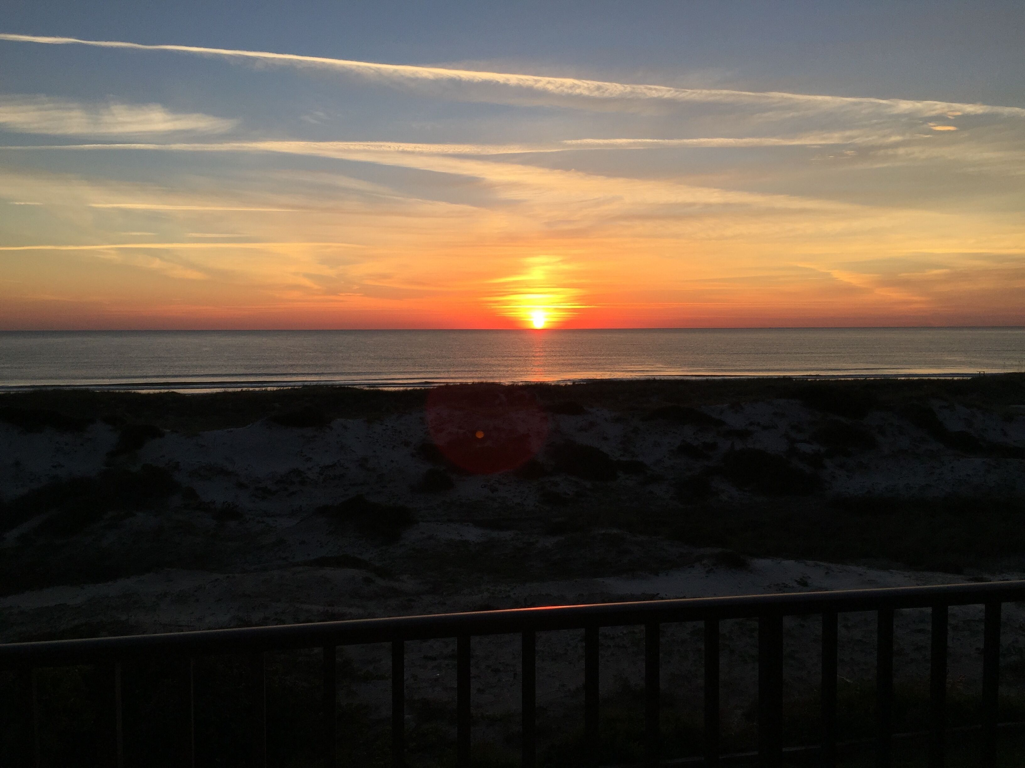 Tramonto su una spiaggia, con una ringhiera in primo piano.
