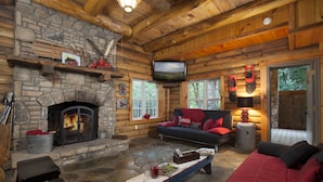 A log cabin interior with a stone fireplace, a television mounted on the wall, and a cozy seating area with a sofa and armchairs.