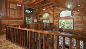 A wooden cabin interior with a balcony, large windows, and a fireplace.