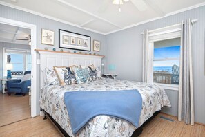 Een slaapkamer met een bed, nachtkastje, stoel, raam met uitzicht en plafondventilator.