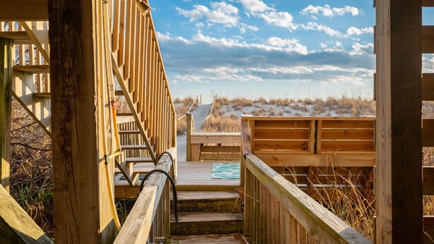 A wooden staircase leading to a deck with a view of a beach and dunes. - Sandy Dunes: Spectacular Oceanfront Home with Amazing Views (Oak Island)