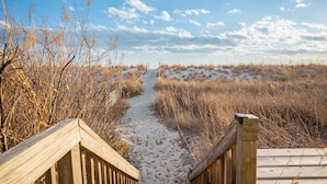 A wooden boardwalk leading through a natural landscape with dry vegetation and a clear sky. - Sandy Dunes: Spectacular Oceanfront Home with Amazing Views (Oak Island)