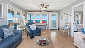 A spacious living room with a view of the ocean, a ceiling fan, and a dining area with a table and chairs. - Sandy Dunes: Spectacular Oceanfront Home with Amazing Views (Oak Island)