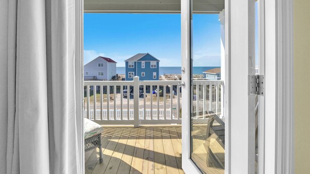 Una terraza de madera con barandal blanco, una silla de mimbre y vista a una playa con casas al fondo.