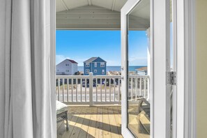 Une terrasse en bois avec une balustrade blanche, une chaise en osier et une vue sur une plage, avec des maisons en arrière-plan.