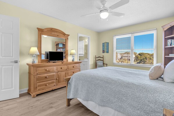 A bedroom with a bed, dresser, television, and a large window with blinds. - Southern Horizons: Elevated Pool, Ocean Views, Near Beach Access (Oak Island)