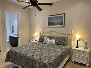 A bedroom with a bed, two bedside tables with lamps, a ceiling fan, a framed picture on the wall, and a bathroom visible through an open door.
