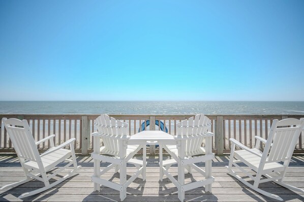 A wooden deck with white chairs and a table, overlooking the ocean.