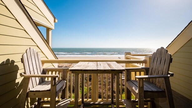Une terrasse en bois avec deux fauteuils Adirondack et une table, donnant sur une plage avec l’océan en toile de fond.