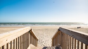 Une promenade en bois mène à une plage d’où l’on a une vue dégagée sur l’océan, et où l’on aperçoit quelques personnes au loin.