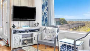 Una habitación con televisión montada en una pared de madera, un armario blanco con cajones, un sillón de mimbre con cojín, y vista al océano a través de una ventana.