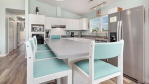 A modern kitchen with a central island, white cabinetry, and teal chairs. - See Otter: Beachwalk cottage in East Beach overlooking the marsh of Davis Canal (Oak Island)
