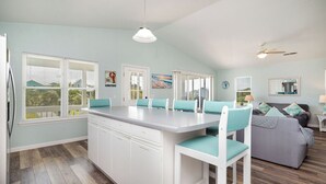 A kitchen with a central island, white cabinetry, and teal chairs. - See Otter: Beachwalk cottage in East Beach overlooking the marsh of Davis Canal (Oak Island)
