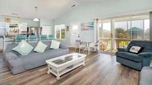 A spacious living area with a grey sofa, a white wicker coffee table, and a blue armchair. - See Otter: Beachwalk cottage in East Beach overlooking the marsh of Davis Canal (Oak Island)