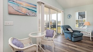 A room with a view of the beach, a wicker chair, a blue armchair, and a small table with a lamp. - See Otter: Beachwalk cottage in East Beach overlooking the marsh of Davis Canal (Oak Island)