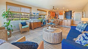 Un vaste espace de vie comprenant un canapé, des fauteuils, une table basse et une table à manger. Il y a également une cuisine équipée de meubles en bois et d’un réfrigérateur. De grandes fenêtres laissent entrer la lumière naturelle et offrent une vue sur l’extérieur.