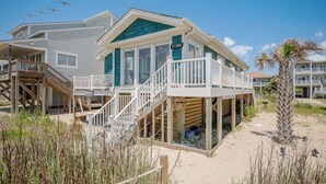Una casa frente a la playa con un porche cubierto, una escalera que lleva a una terraza y una zona de arena.