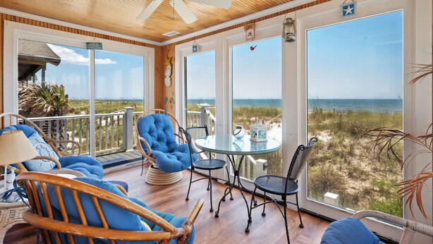 Una habitación iluminada por el sol con techo de madera, ventanas amplias con vistas a la playa y una mesa de vidrio con sillas.