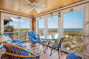 Una habitación iluminada por el sol con techo de madera, ventanas amplias con vistas a la playa y una mesa de vidrio con sillas.