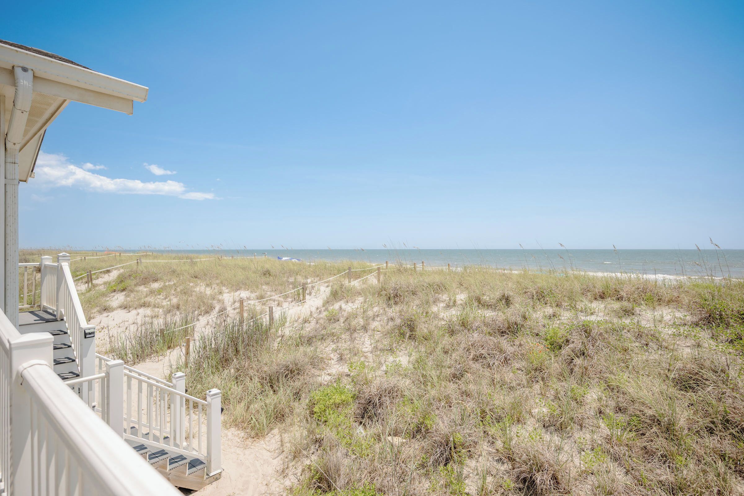 A beach view from a white building with a wooden deck and railing, leading to a sandy path.