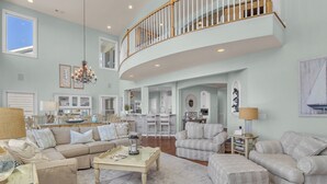 A spacious living area with a high ceiling, a staircase leading to an upper level, and a view of the kitchen and dining area. - Surviving Stress: Exquisite Oceanfront Caswell Beach Home Perfect for Small Beac (Caswell Beach)