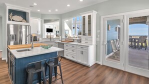 A modern kitchen with a central island, white cabinetry, and a view to an outdoor deck. - Surviving Stress: Exquisite Oceanfront Caswell Beach Home Perfect for Small Beac (Caswell Beach)