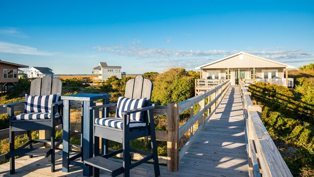 A wooden deck with two Adirondack chairs and a table, leading to a building with a white facade and large windows. - Caswell Cabana: Oceanfront, Split Floor Plan, Views of Lighthouse (Caswell Beach)