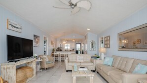 A spacious living room with a television, comfortable seating, and a kitchen in the background. - Caswell Cabana: Oceanfront, Split Floor Plan, Views of Lighthouse (Caswell Beach)