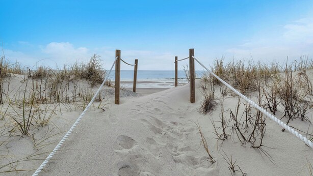 Ein Sandweg am Strand, der zum Meer führt, gesäumt von Holzpfählen und Seilen.