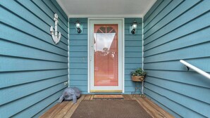 A wooden porch with a red-framed door, a small gray cat statue, and a potted plant. - Hook Wine and Sinker: Fenced Yard for Dogs, Hot Tub! (Yaupon Beach)