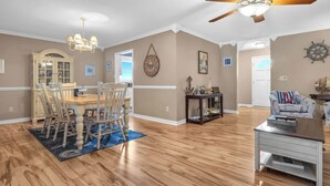 A spacious living room with hardwood flooring, a dining area with a wooden table and chairs, a ceiling fan, and a view of the outside. - Hook Wine and Sinker: Fenced Yard for Dogs, Hot Tub! (Yaupon Beach)