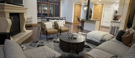 A spacious living room with a large sofa, a coffee table, and a fireplace. There is a dining area with chairs and a table, and a kitchen area with cabinets and appliances.