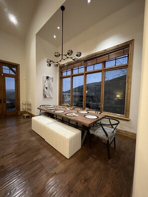 Enjoy meals in this dining area with a long table, high ceiling, and large windows offering a view of the outdoors.