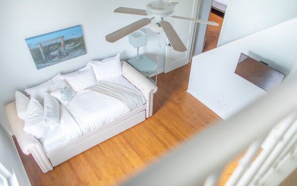 Et soveværelse med en hvid seng, ventilator i loftet, et natbord og et billede i ramme på væggen.