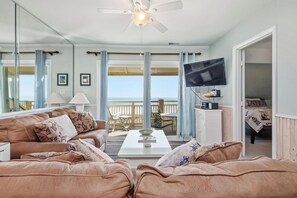Een woonkamer met uitzicht op de oceaan, een plafondventilator, een flatscreen televisie en een eethoek met een tafel en stoelen.