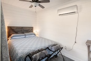 A bedroom with a bed, a ceiling fan, an air conditioning unit, and a bedside table.
