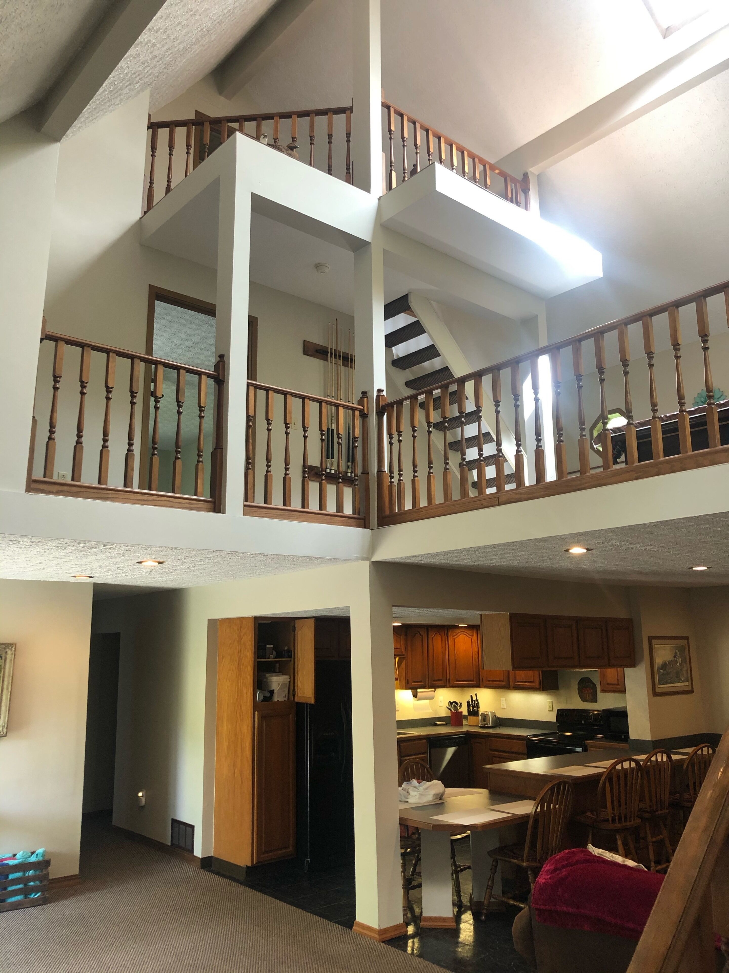 Un intérieur à plusieurs niveaux, avec un escalier reliant deux étages, des rampes en bois, un espace cuisine doté de meubles et d’appareils électroménagers, et un coin repas avec des chaises et une table.