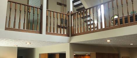 A multi-level interior with a staircase connecting two floors, wooden railings, a kitchen area with cabinets and appliances, and a dining area with chairs and a table.