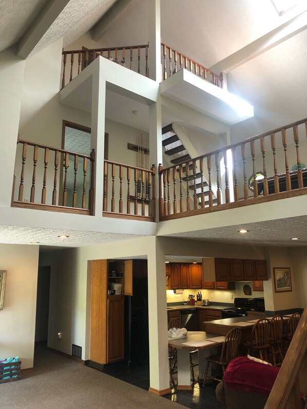 A multi-level interior with a staircase connecting two floors, wooden railings, a kitchen area with cabinets and appliances, and a dining area with chairs and a table. - Peaceful Hocking Hills Vacation Retreat (Rockbridge)