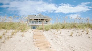 Une maison de plage dotée d’une terrasse en bois menant à l’entrée, entourée de sable et d’herbes hautes.