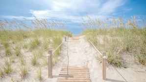 Un sentier en bois mène à une plage de sable, de chaque côté bordée d’herbe.