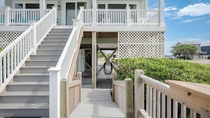 Un escalier en bois mène à un porche abrité, doté d’une balustrade blanche et d’une clôture à treillis, avec un ciel dégagé en arrière-plan.