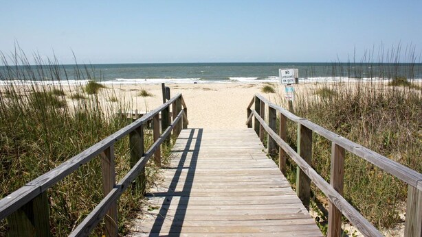 Una passerella in legno che conduce a una spiaggia, con una postazione di salvataggio visibile in lontananza.