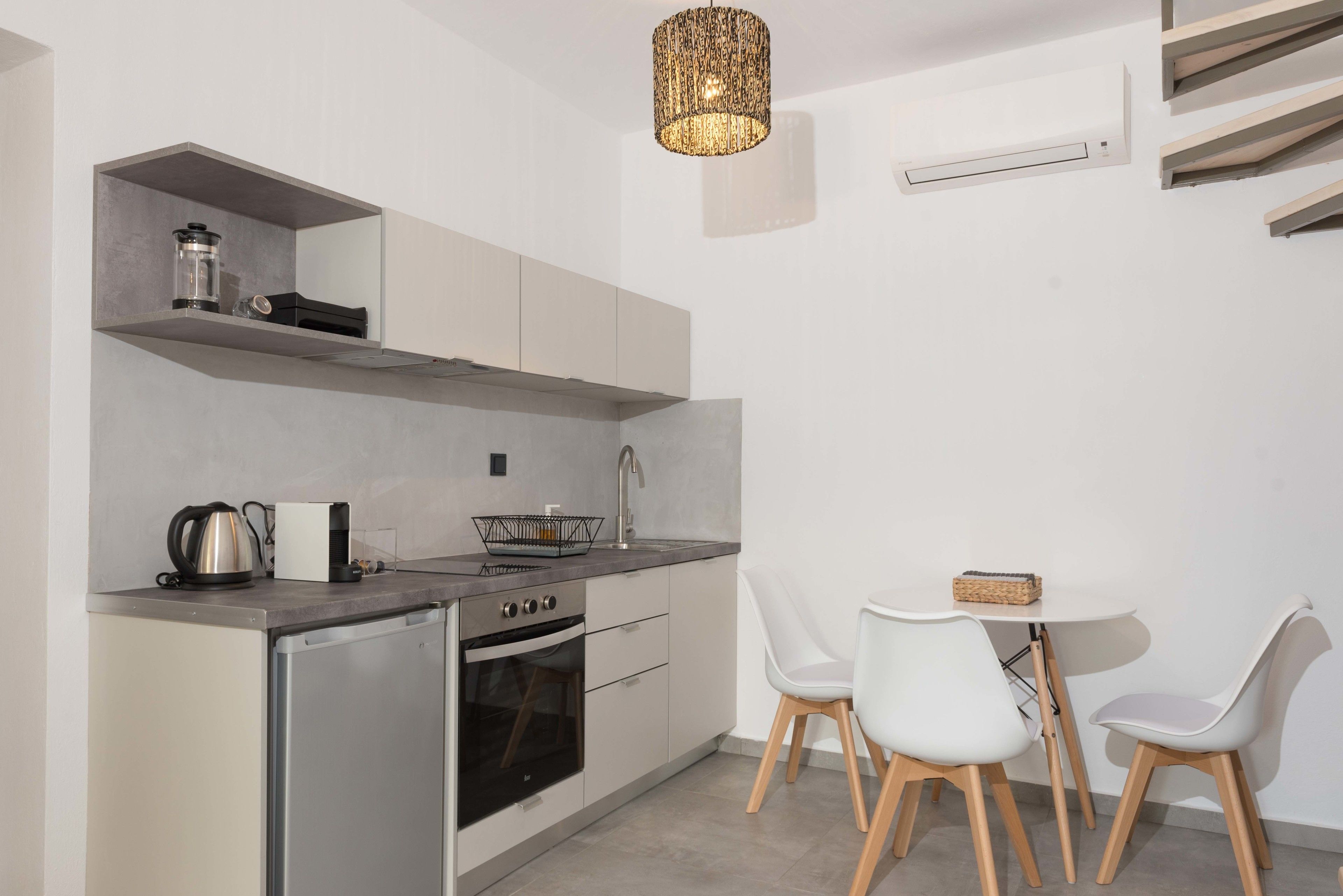 A modern kitchen with a built-in oven, a coffee maker, and a kettle on the countertop. There is a dining table with white chairs and a basket on it.