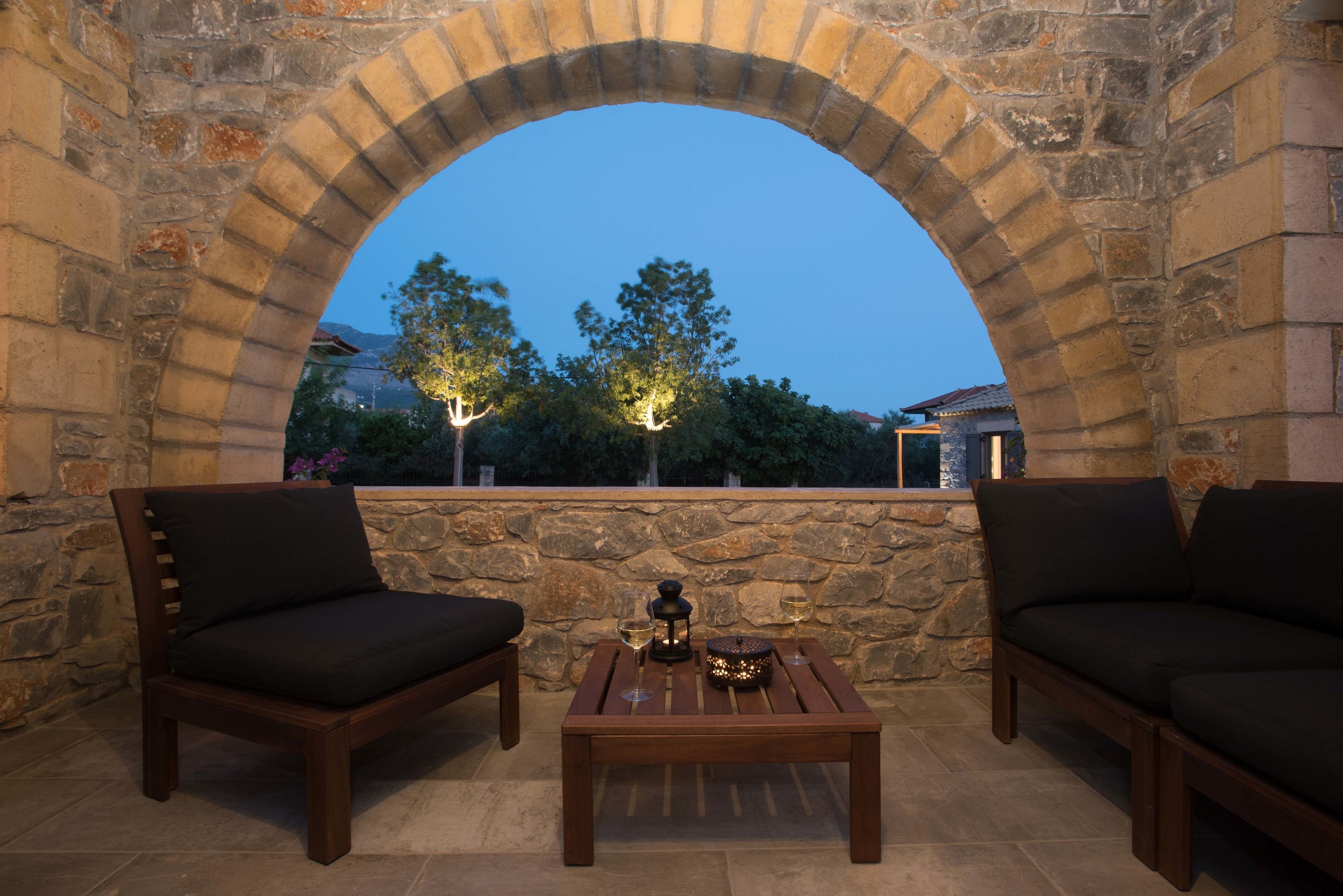 An outdoor seating area with stone walls and a wooden table, featuring two cushioned chairs and a lantern.