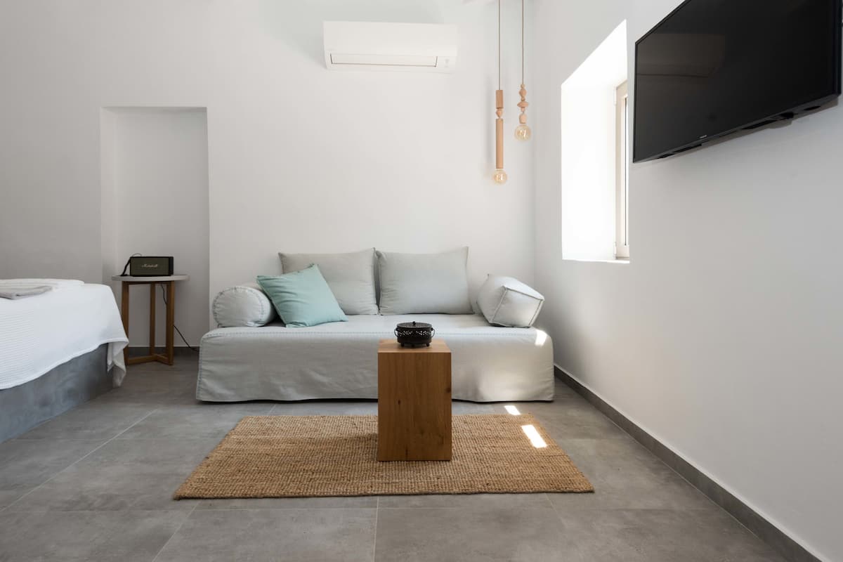 Un dormitorio moderno y minimalista con cama, sofá, mesita de madera, televisor y ventana que deja entrar luz natural.