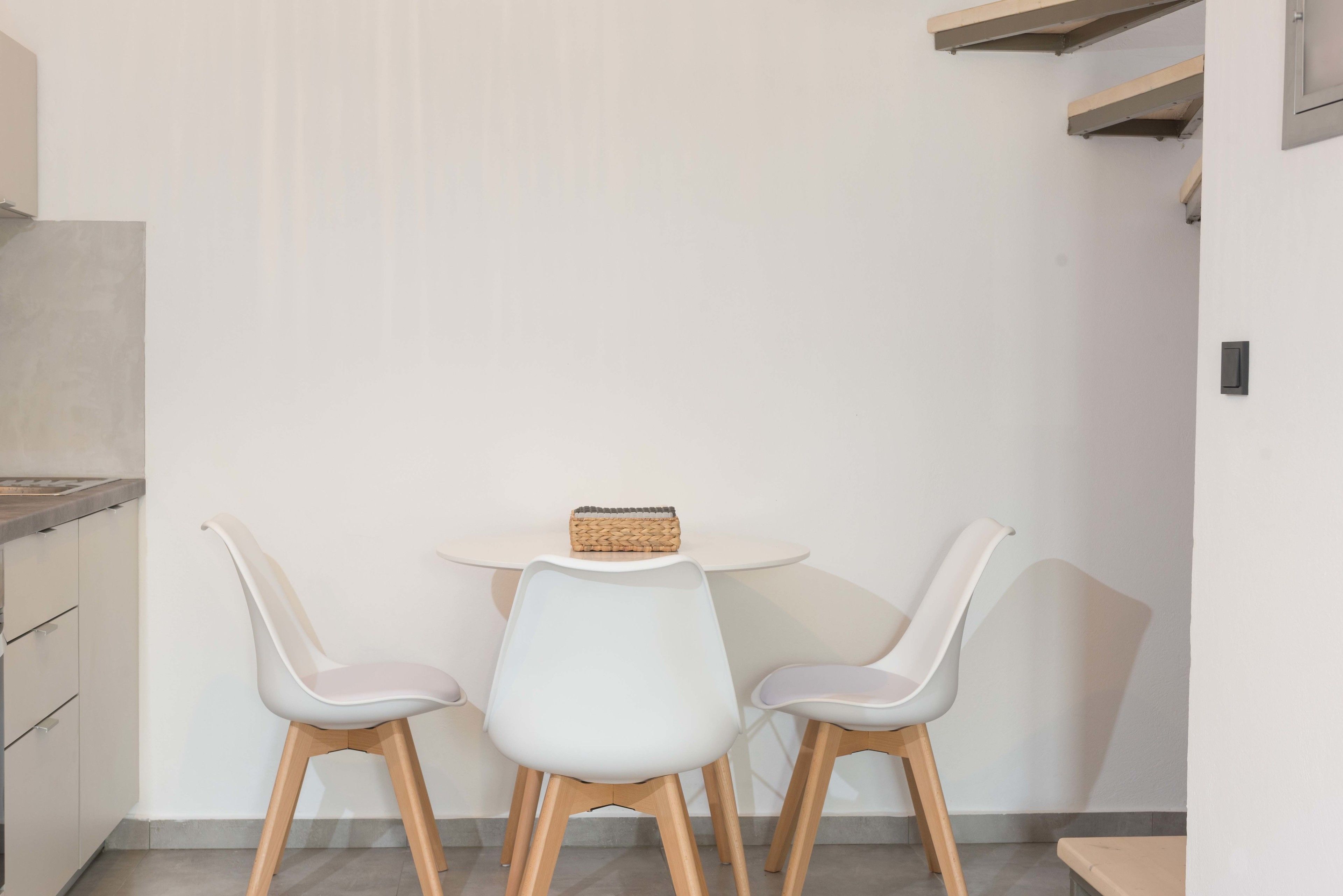 A small, modern kitchen with white cabinets, a round table, and two white chairs with wooden legs.