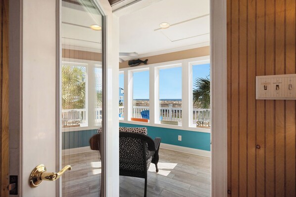 A view through a glass door into a room with a wooden floor, a patterned chair, and a view of the outside deck.