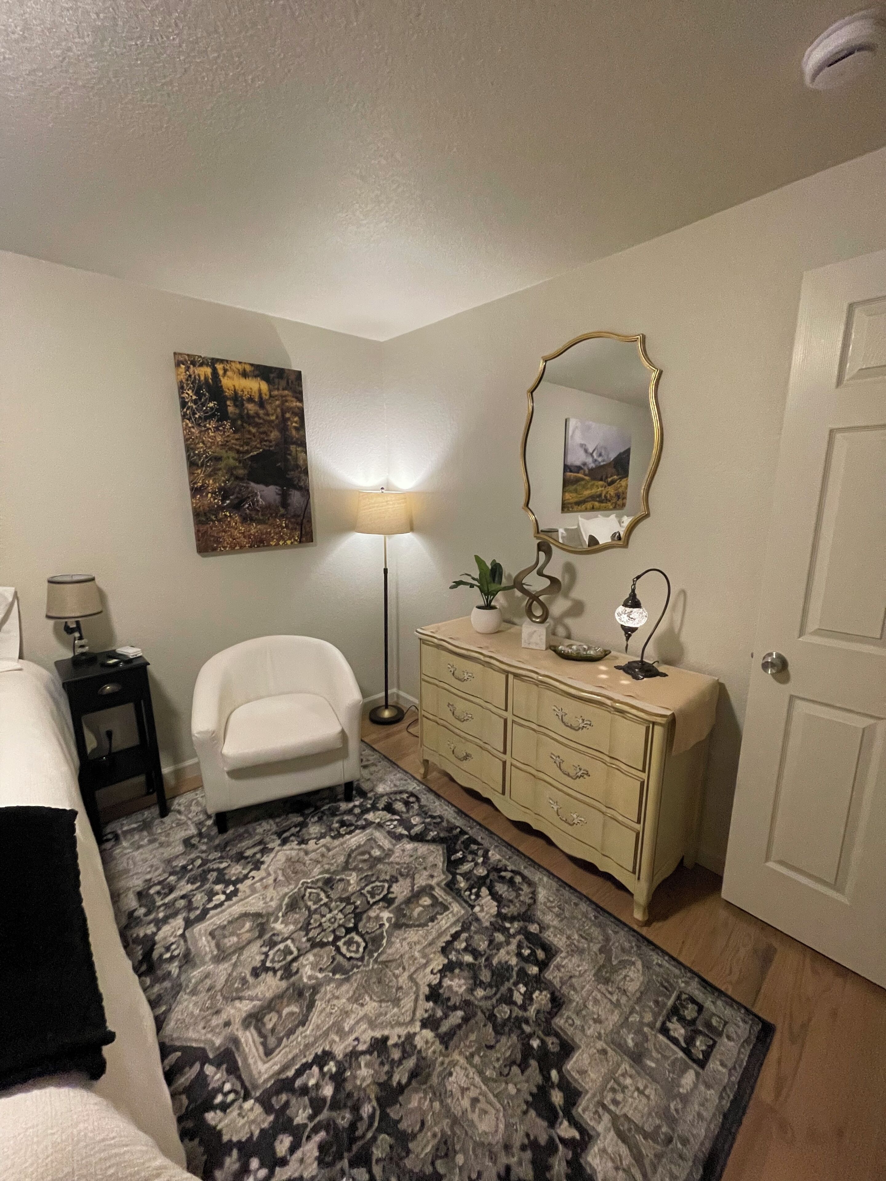 A bedroom with a bed, a white armchair, a dresser, a mirror, a lamp, and a patterned rug.