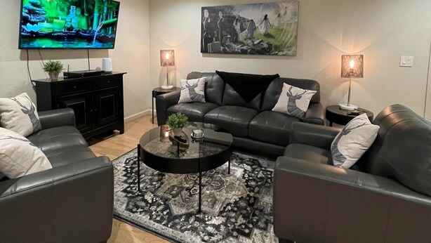 A modern living room with a black leather sofa, a glass coffee table, and a large flat-screen TV mounted on the wall. - Fantastic Denver Location! 5 mins from every major highway! Mountain & Lake View (Denver)